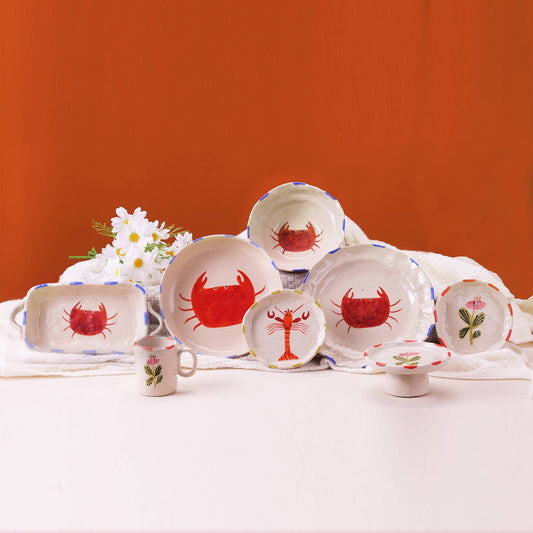 Set of ceramic plates and cups with red crab designs on a white tablecloth against an orange wall.