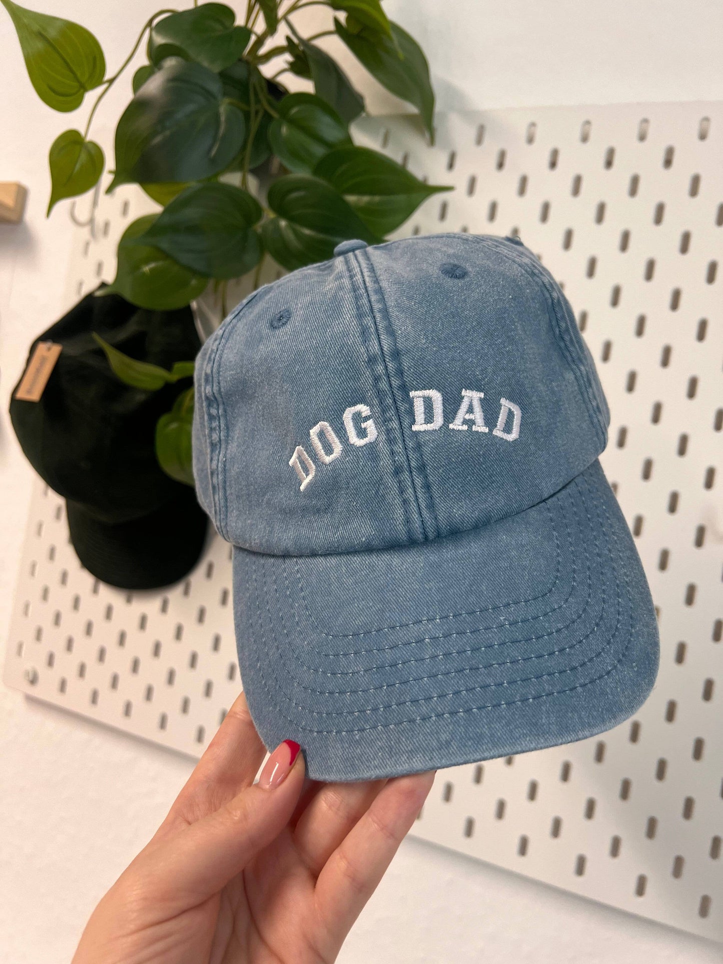 Blue cap with 'DOG DAD' text held by a hand against a white pegboard background.