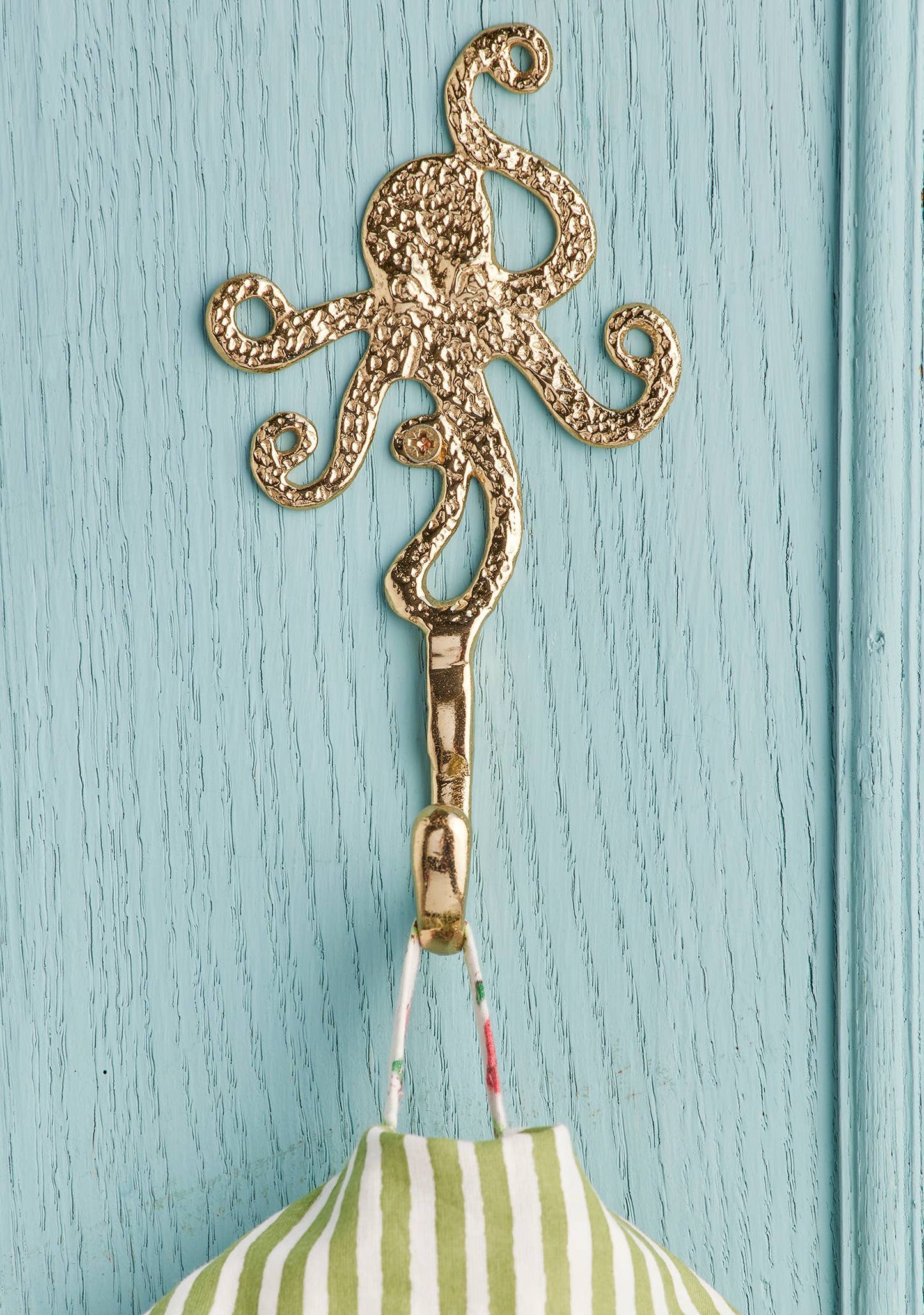 A gold octopus-shaped hook hanging on a light blue painted wooden door, with a green and white striped fabric item suspended from it.