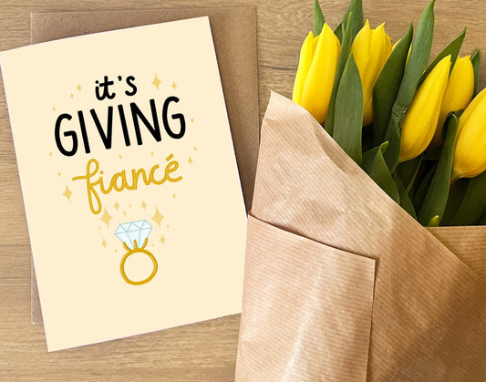 Greeting card with 'it's GIVING fiancé' text and yellow tulips in a brown envelope on a wooden surface.