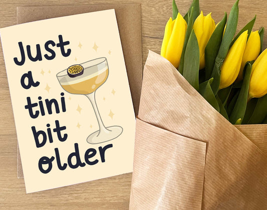 Greeting card with a cocktail illustration and text, next to a bouquet of yellow tulips in an envelope on a wooden surface.