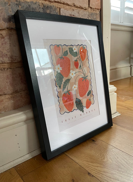 A framed art print featuring a hand-drawn illustration of strawberries and a fruit market backdrop, placed on a floor surface.