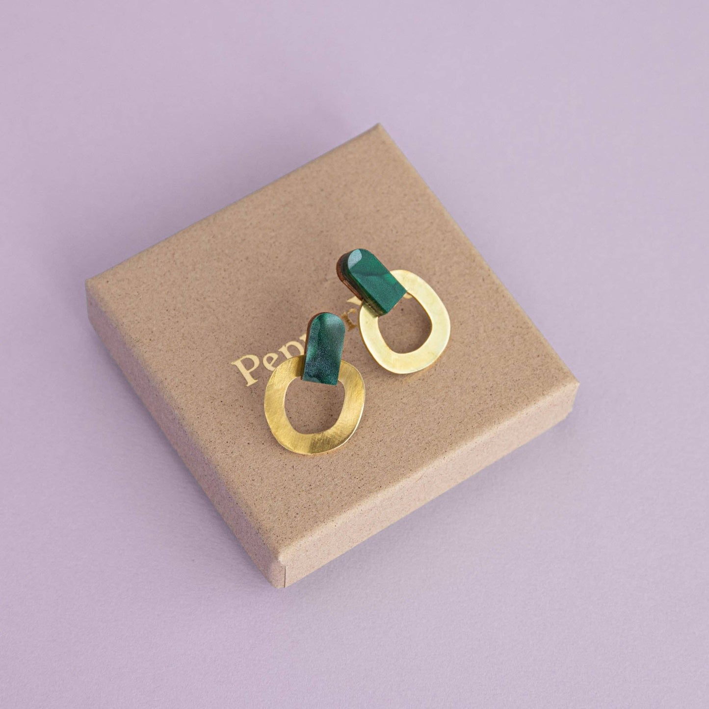 Gold and green earrings on a brown jewelry box with 'Pepper You' branding on a purple background.
