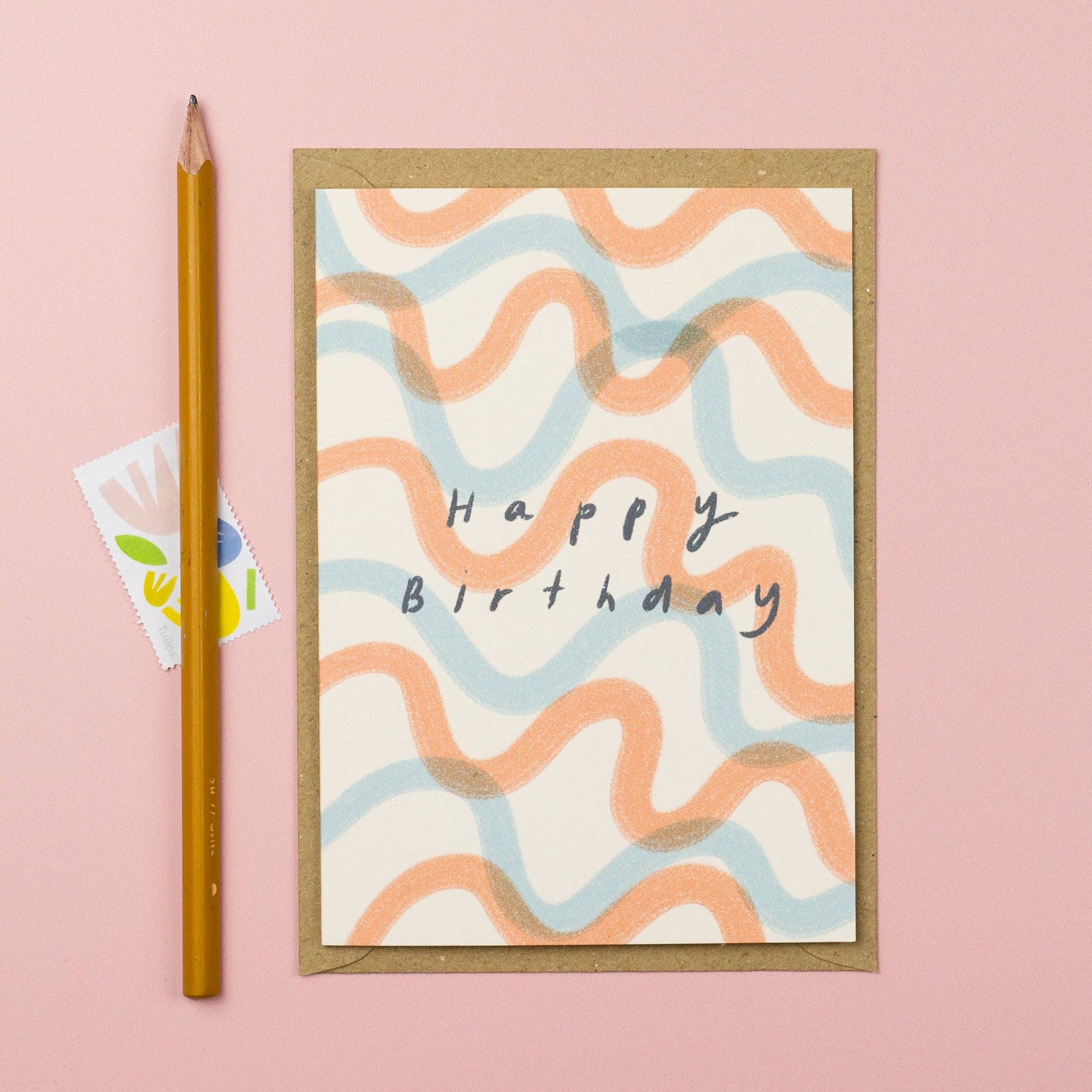 Birthday card with wavy patterns and 'Happy Birthday' text on a pink background, accompanied by a pencil and small card.