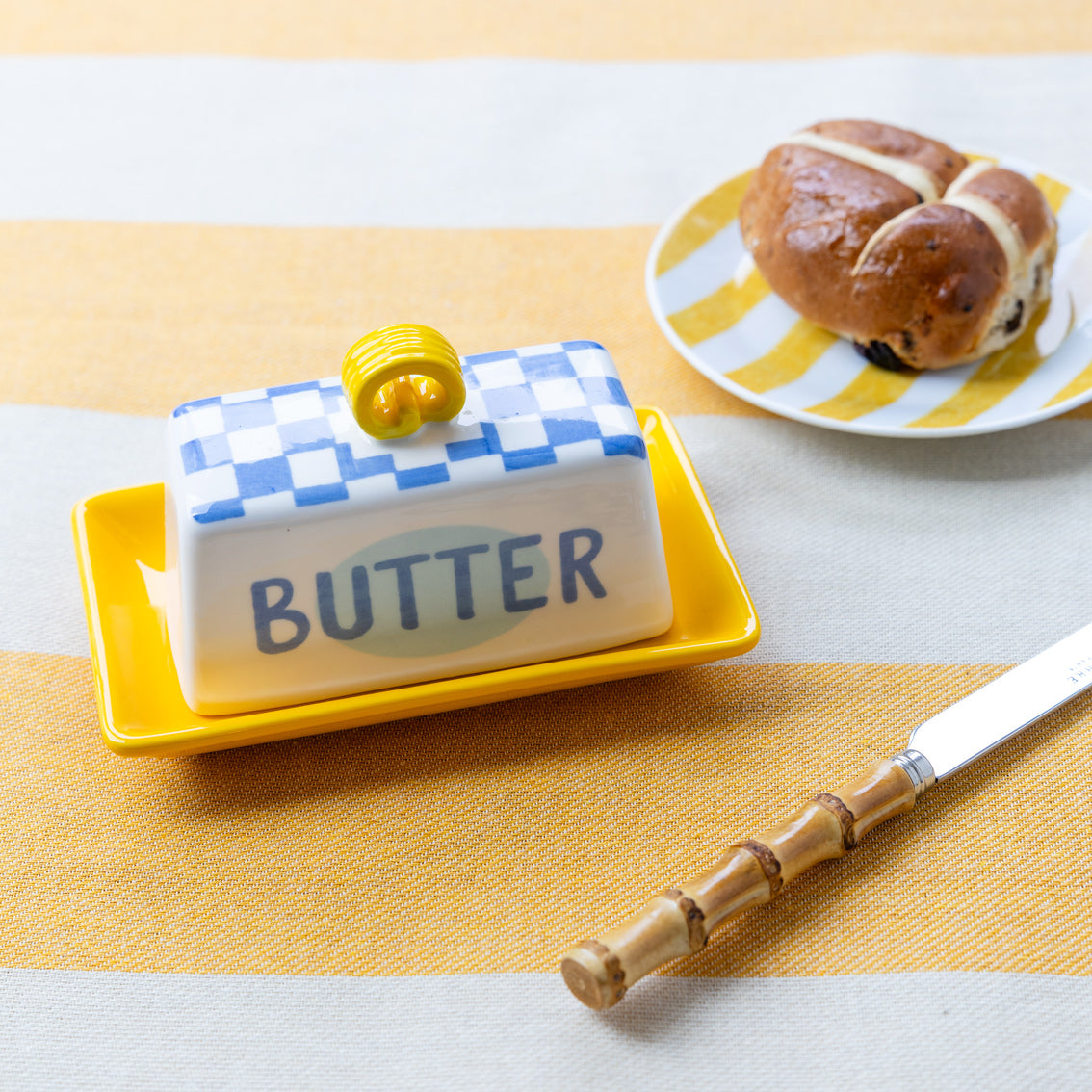 Butter dish with checkered lid on a yellow tray, next to a plate of pastries.