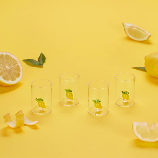 Four clear cylindrical cups arranged in a row on a bright yellow surface, each containing a small lemon illustration printed near the bottom. Lemon slices, peels, and a whole lemon are placed around the cups as styled props.