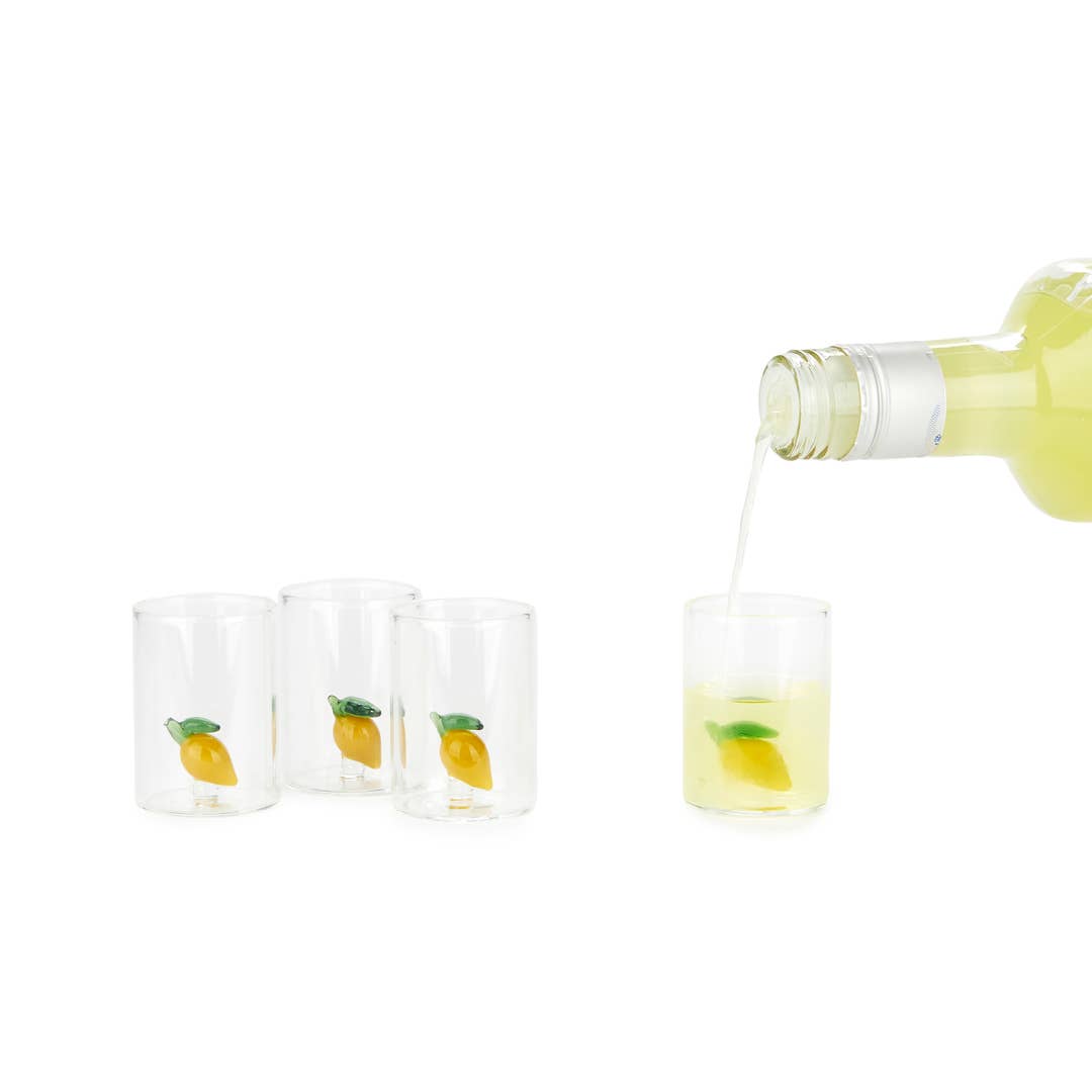 Four clear shot glasses with small yellow lemon illustrations, shown on a white background with a bottle pouring pale yellow liquid into one glass.