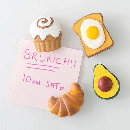 Decorative magnets shaped like a croissant, avocado, toast with an egg, and a cupcake on a white background.