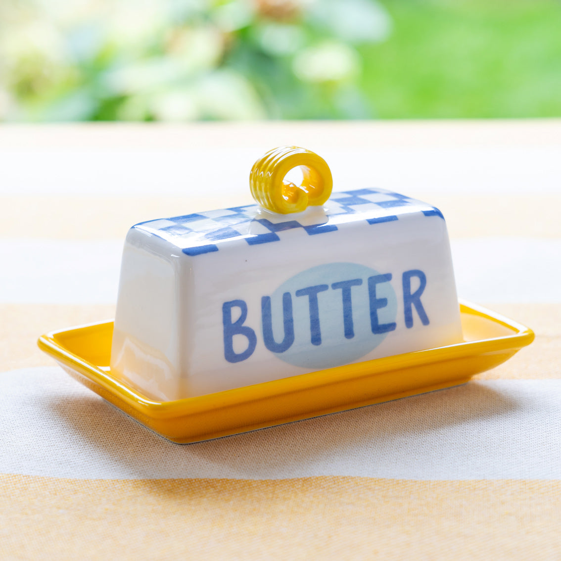 Butter dish with blue lid and yellow handle on a yellow tray, set against a blurred green outdoor background.