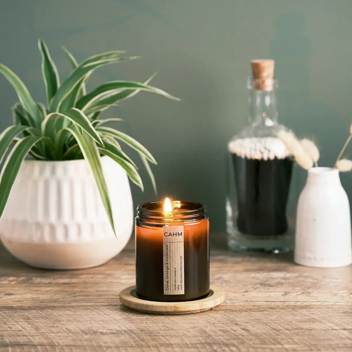 Candle with a label on a wooden surface next to a plant and decorative bottles.