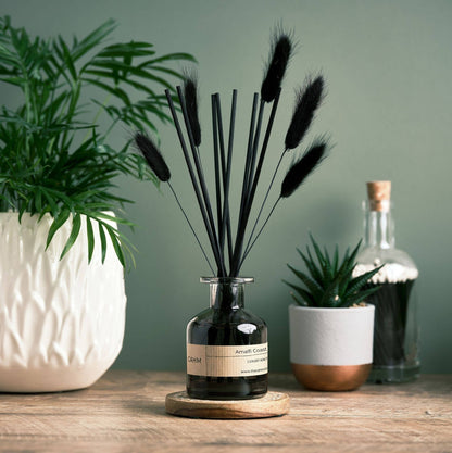 Decorative diffuser with black dried grasses in a glass bottle on a wooden surface with plants in the background.