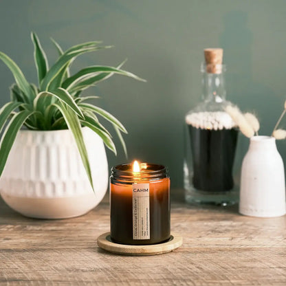Candle with a label on a wooden surface next to a plant and decorative bottles.