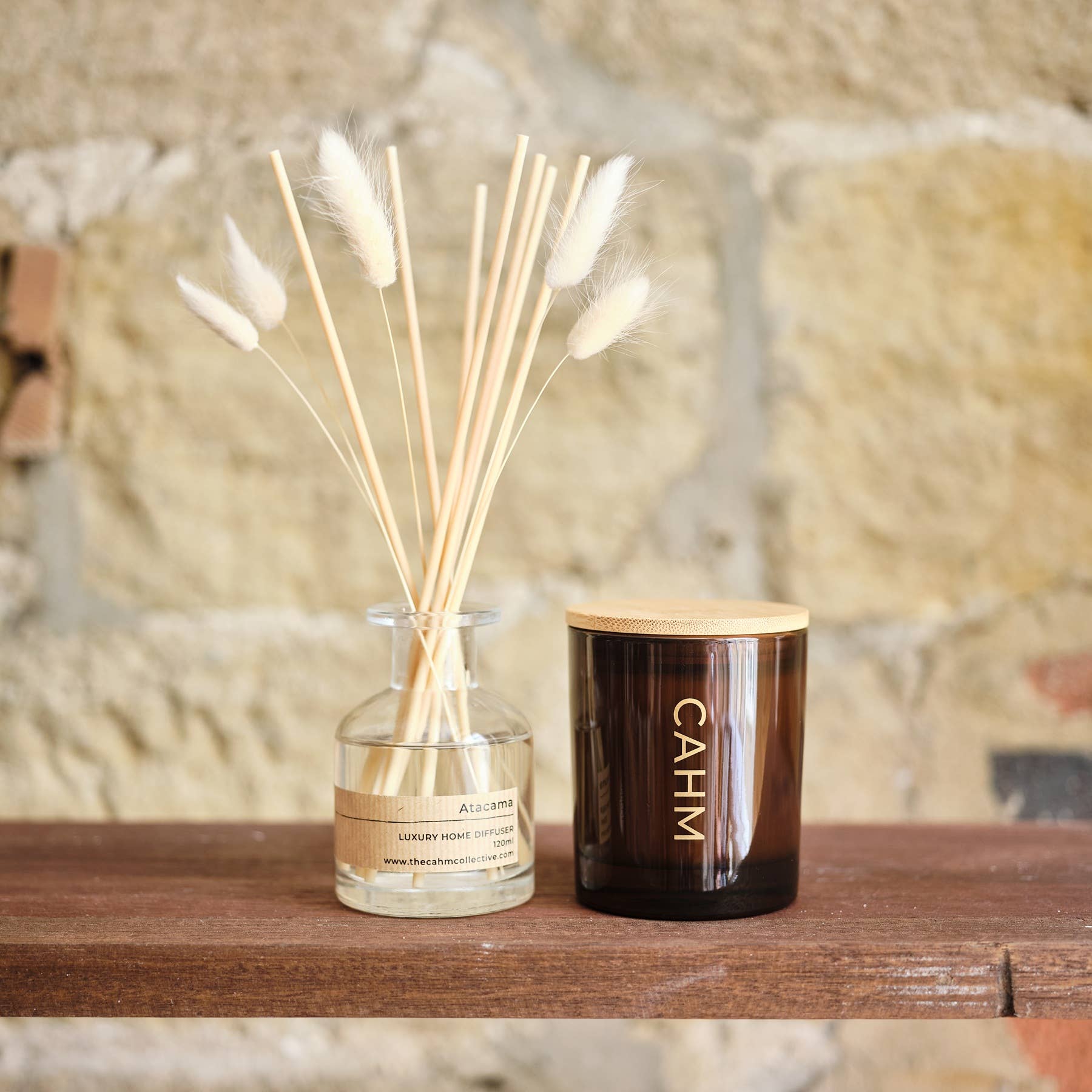 A reed diffuser and a candle with a wooden lid on a wooden surface against a stone wall background.