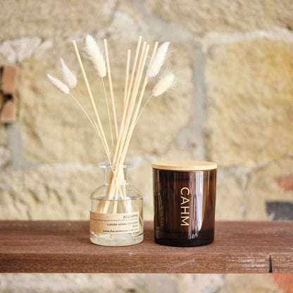 A reed diffuser and a candle with a wooden lid on a wooden surface against a stone wall background.
