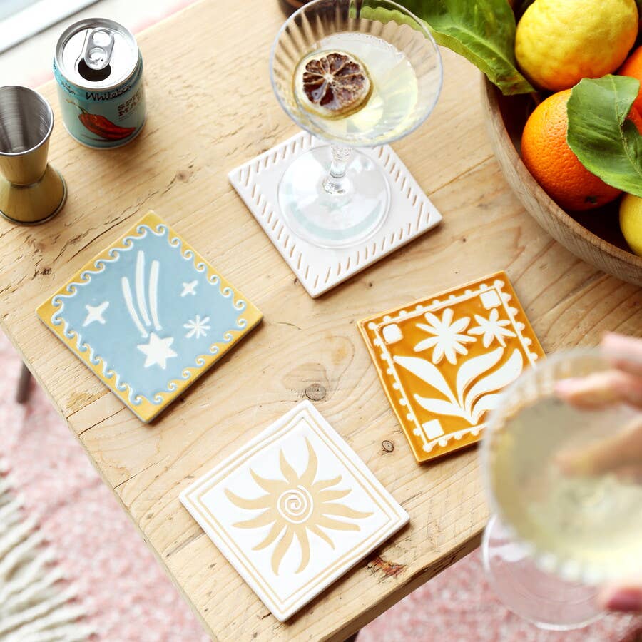 Coasters with geometric designs on a wooden table with drinks and fruit.