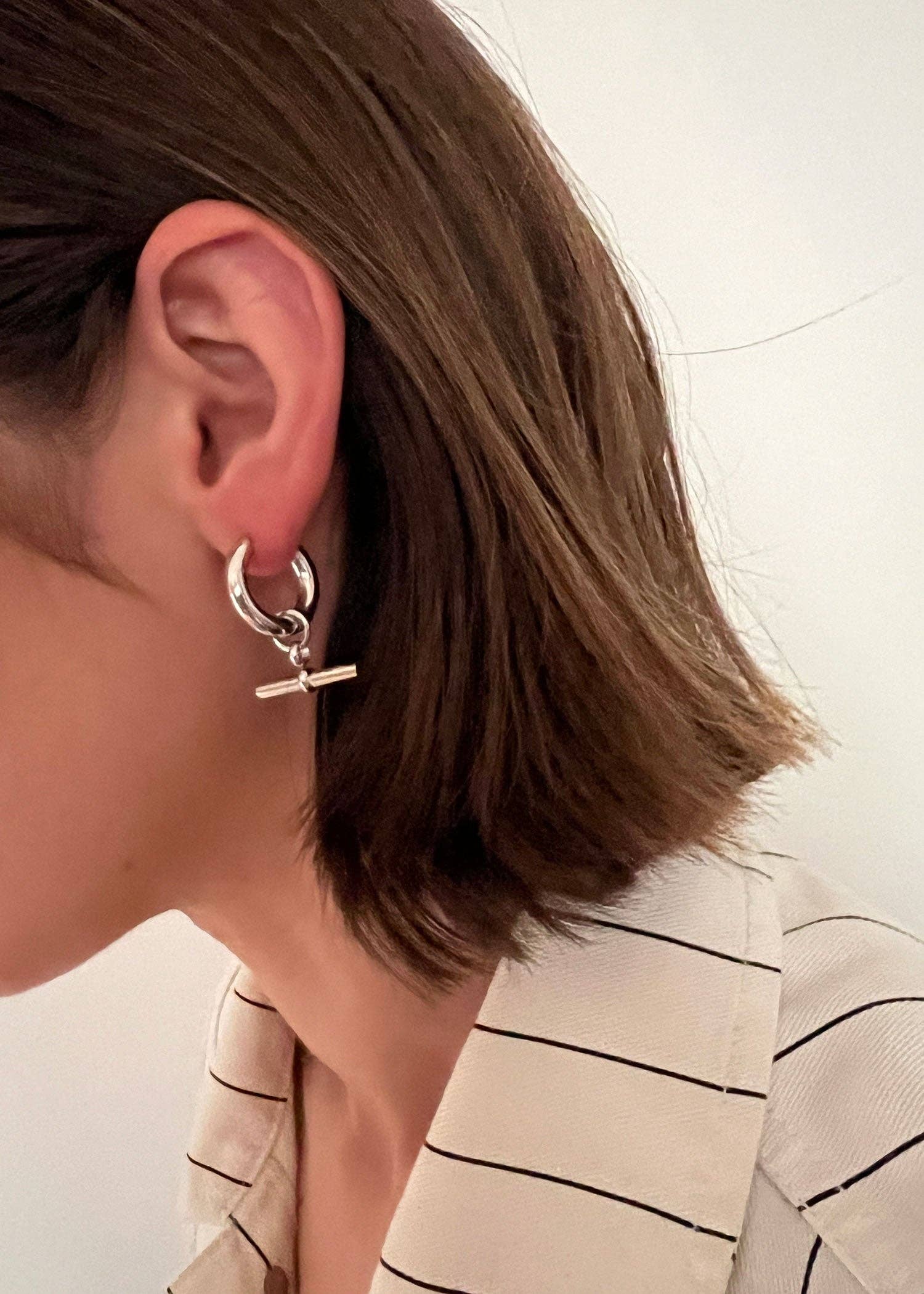 Close-up of an ear wearing a silver hoop earring with a small pendant, against a neutral background.