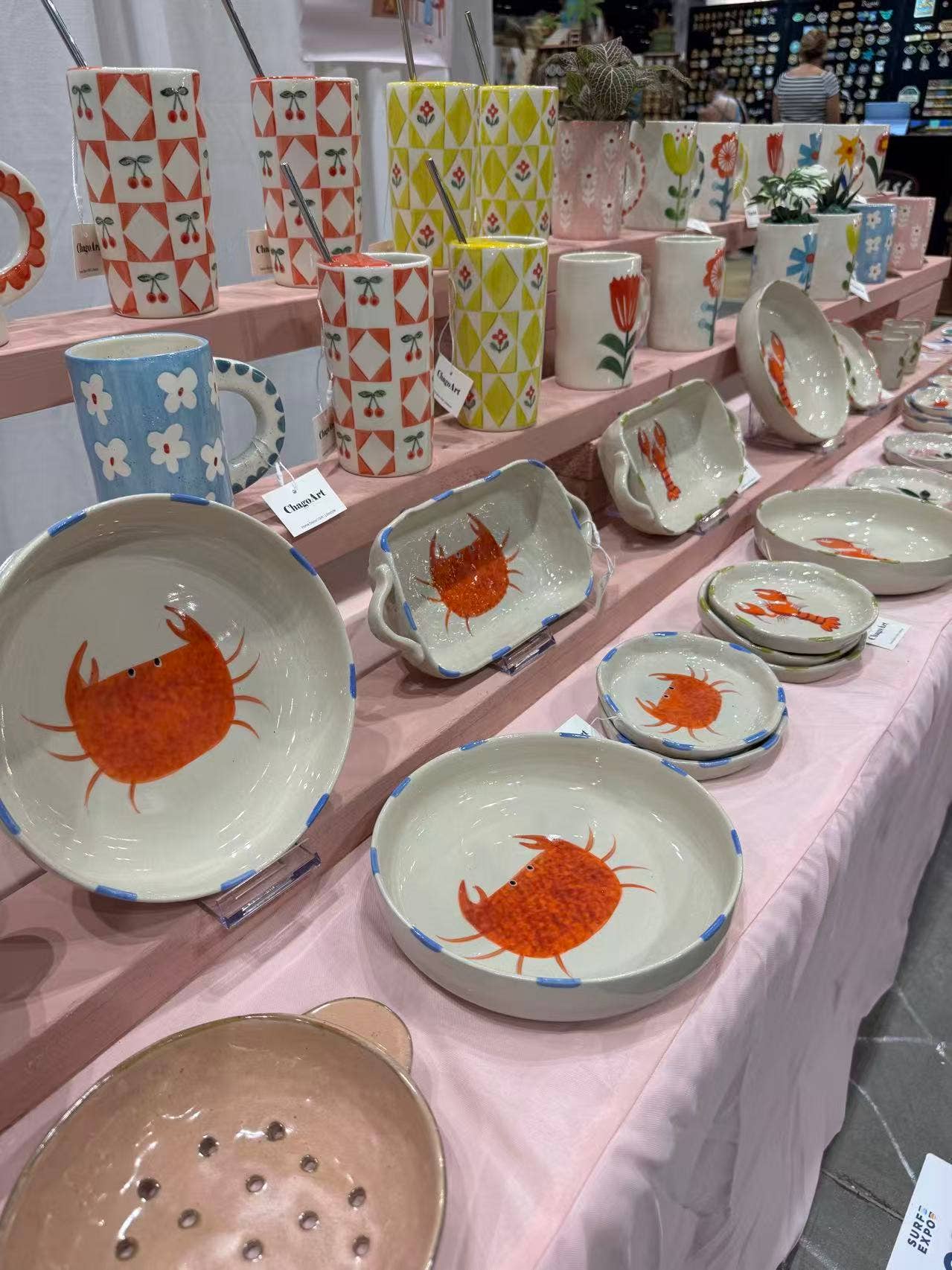 Display of ceramic plates and mugs with crab design on a pink tablecloth.