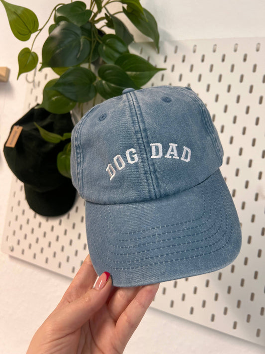 Blue cap with 'DOG DAD' text held by a hand against a white pegboard background.