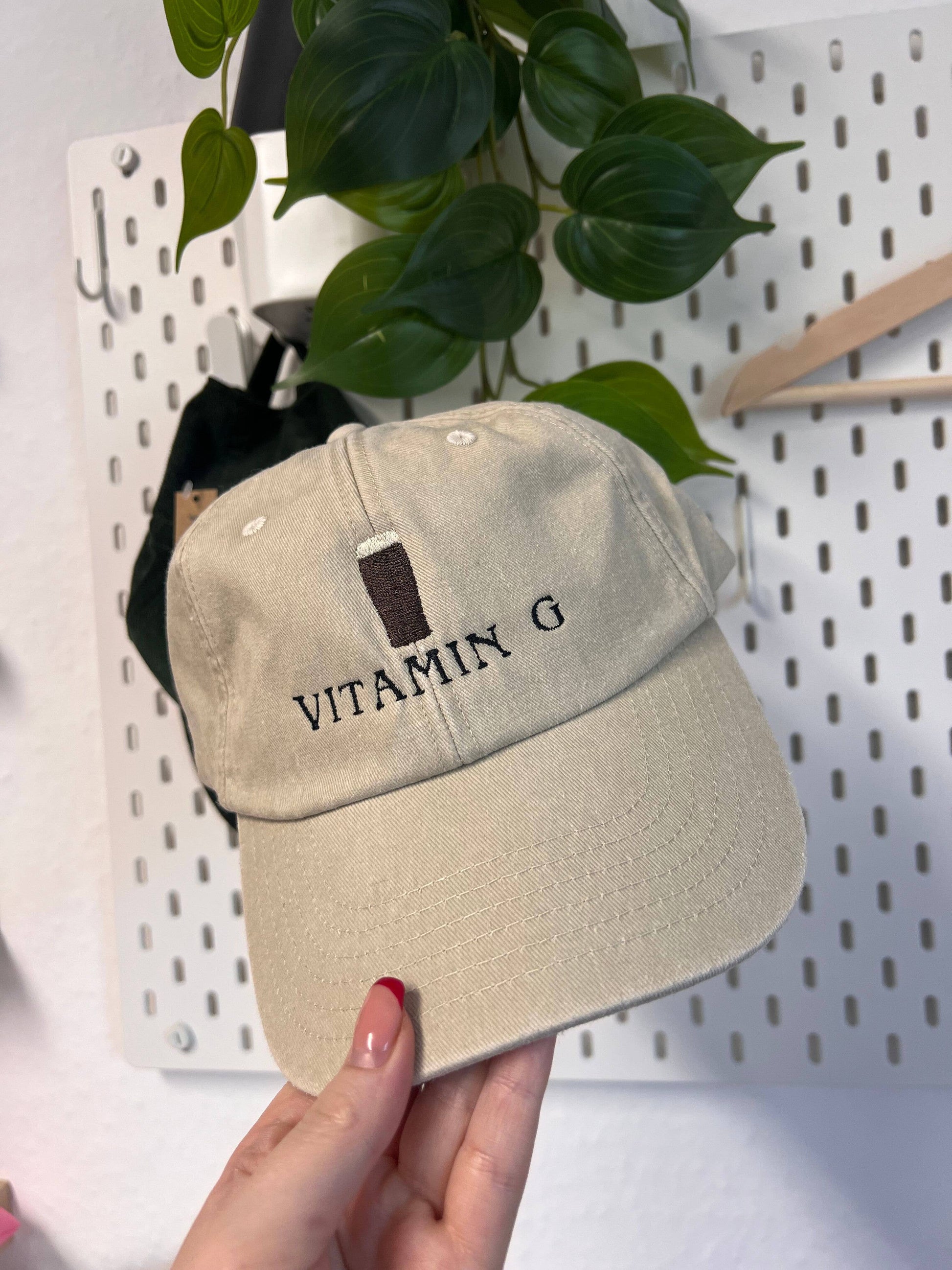Beige cap with 'Vitamin G' text held by a hand against a white pegboard background