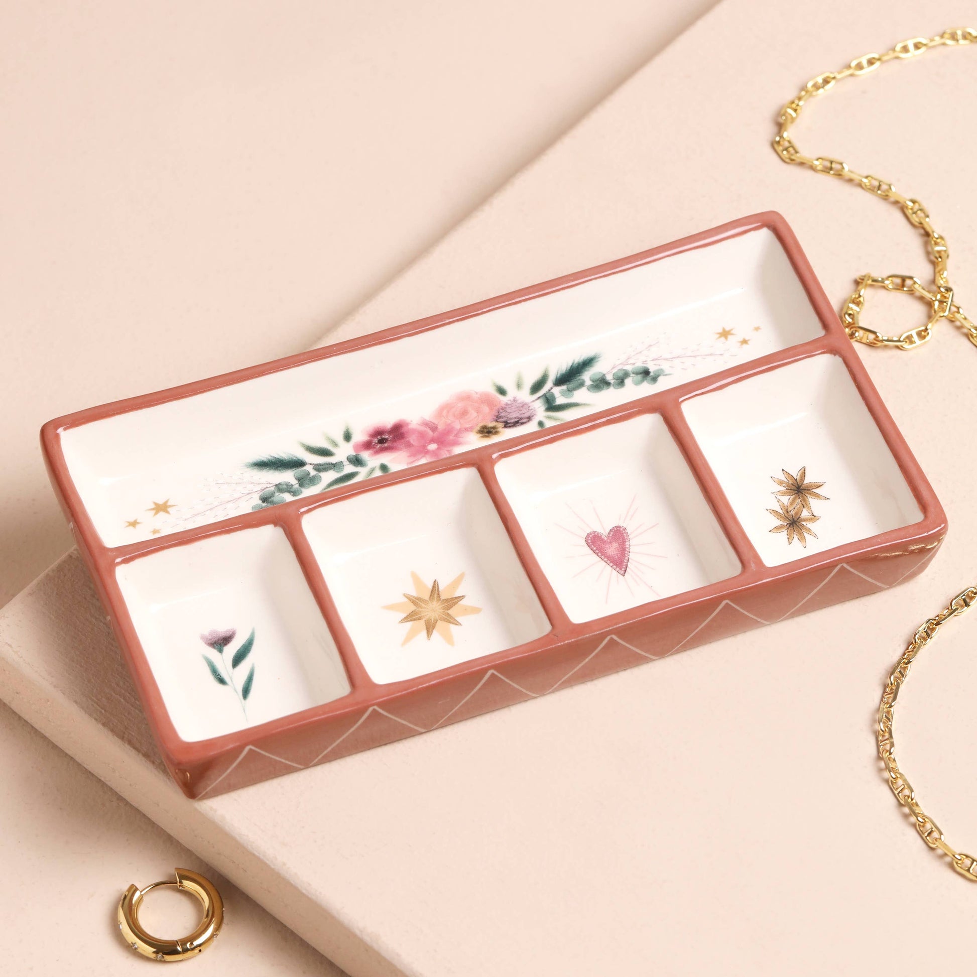 A floral ceramic trinket tray with four square compartments and one rectangle compartment, displayed on a neutral background, with a gold-colored necklace placed next to it.