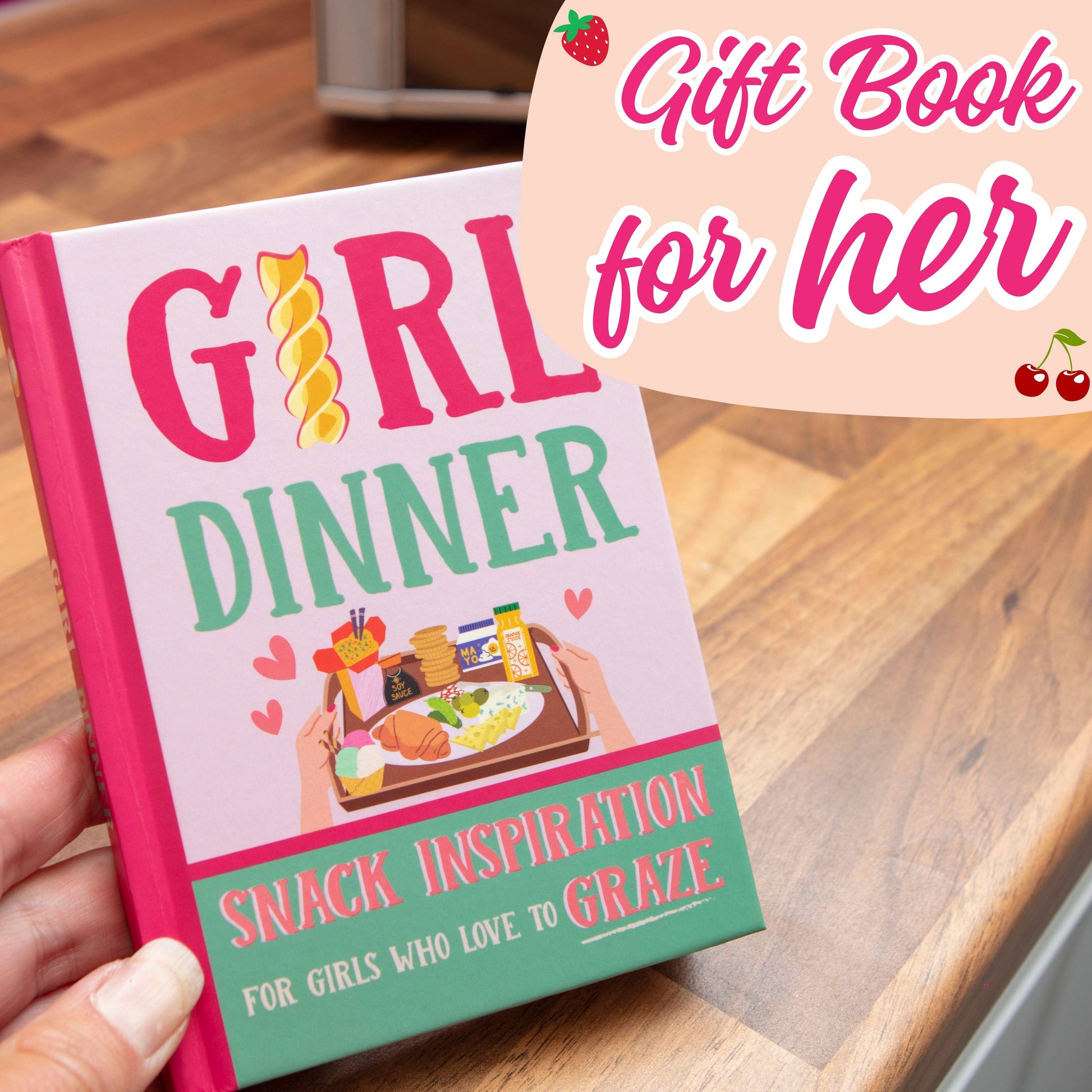 Book titled 'Girl Dinner' held by a hand on a wooden surface with promotional text overlay.