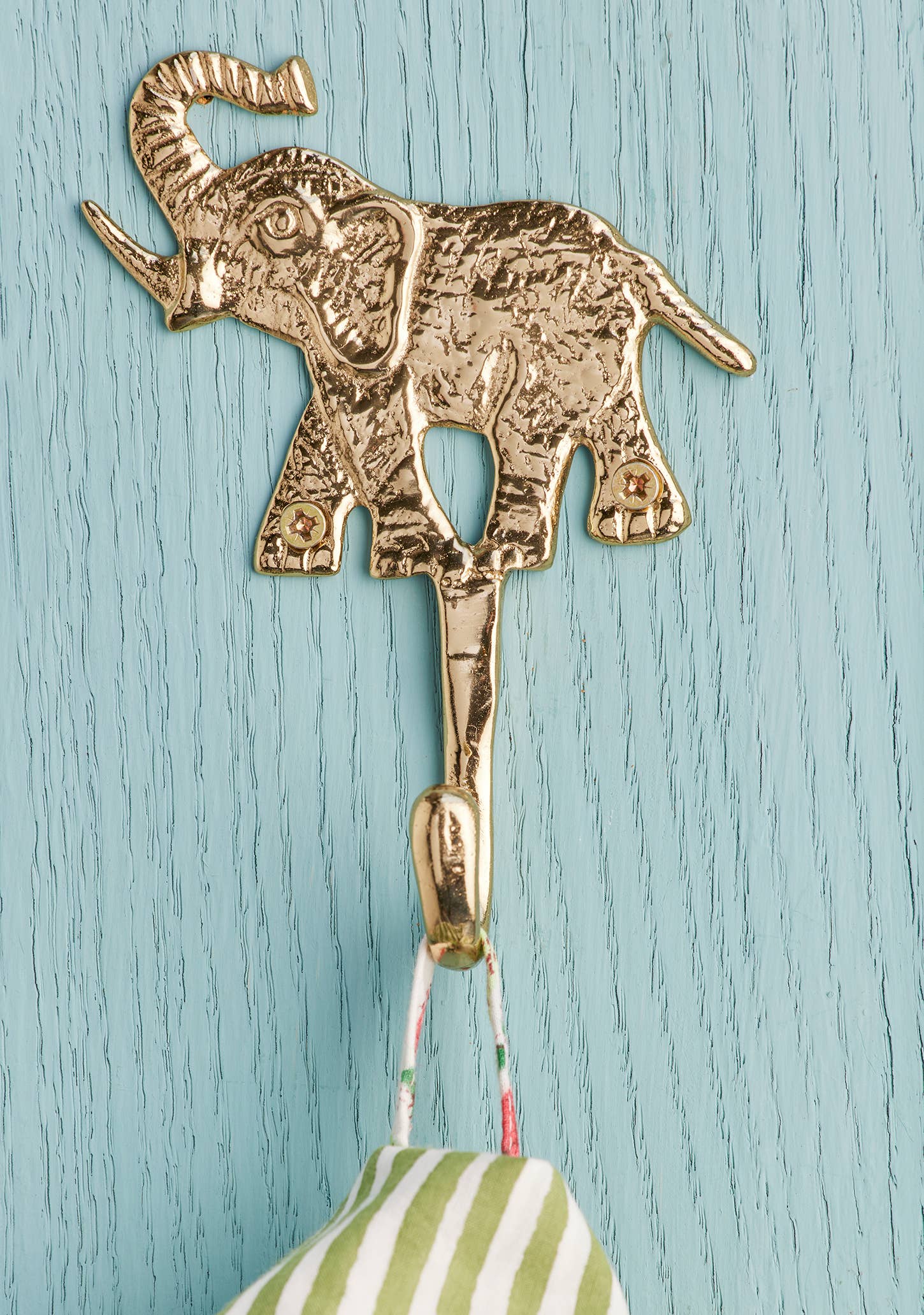 A gold elephant-shaped hook with a textured finish, mounted on a light blue painted wood wall. A green and white striped fabric item hangs from the hook.