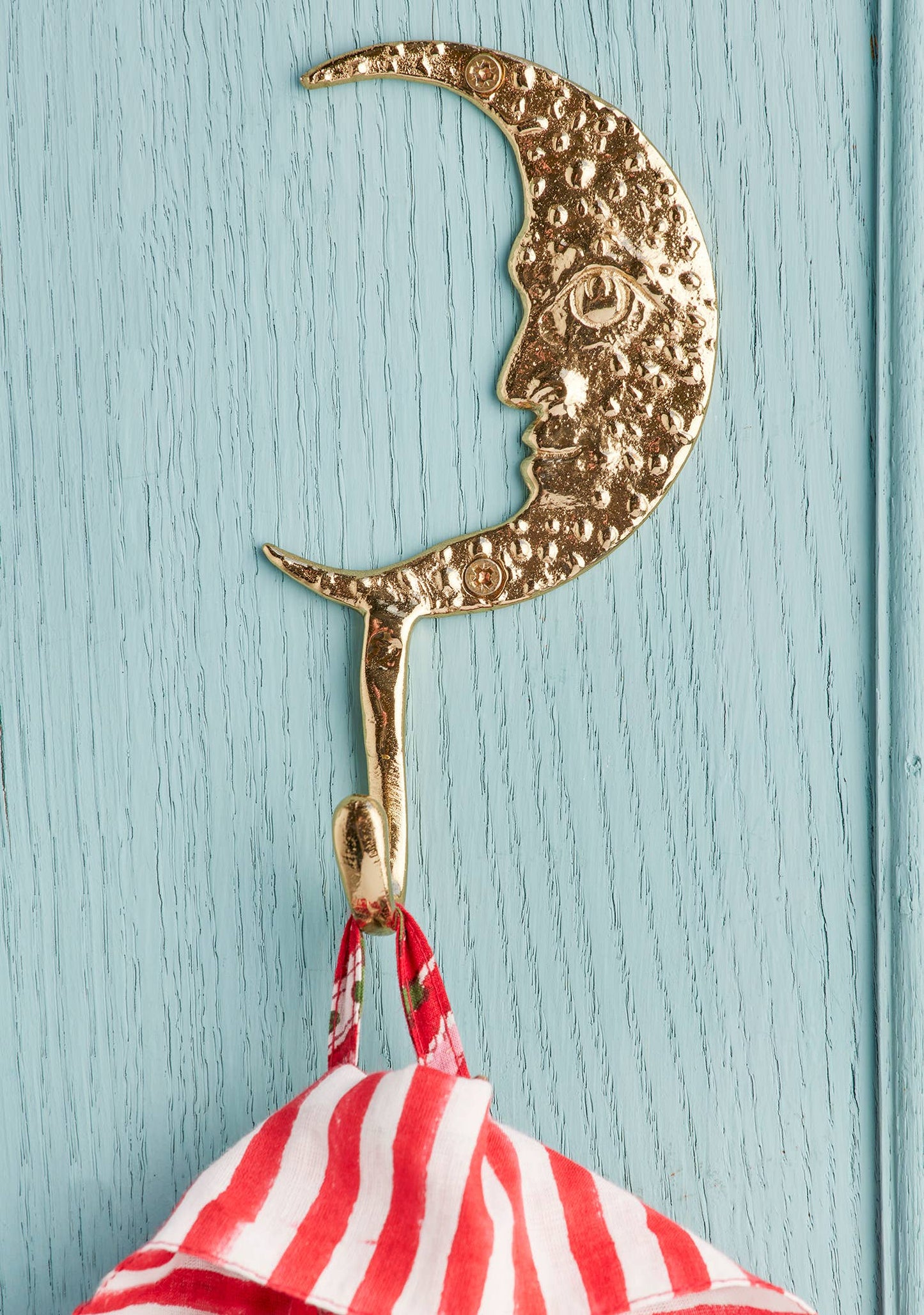 A gold crescent moon wall hook with a face design hanging on a light blue painted wooden door, with a red and white striped fabric item hanging from it.