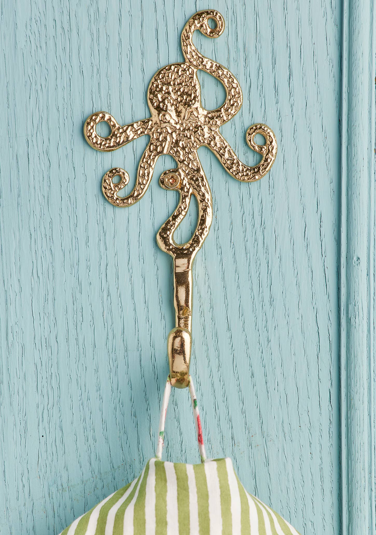 A gold octopus-shaped hook hanging on a light blue painted wooden door, with a green and white striped fabric item suspended from it.