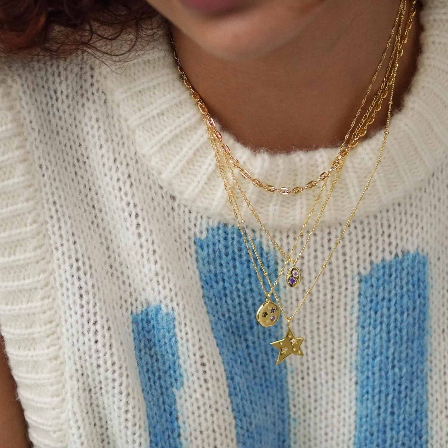 Close-up of a person wearing gold necklaces with star and round pendants on a white sweater with blue stripes.
