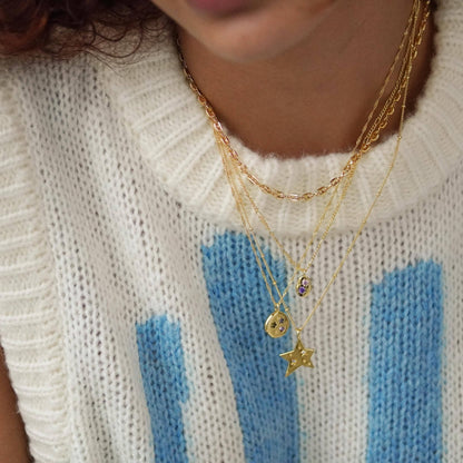 Close-up of a person wearing gold necklaces with star and round pendants on a white sweater with blue stripes.