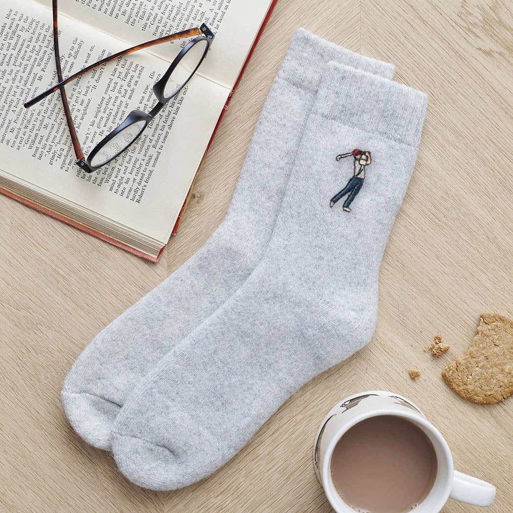 Gray socks with a small embroidered design on a wooden surface with a book, glasses, and a mug.