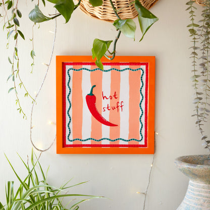 Decorative wall art with a red chili pepper and 'hot stuff' text on a striped background, framed in orange.