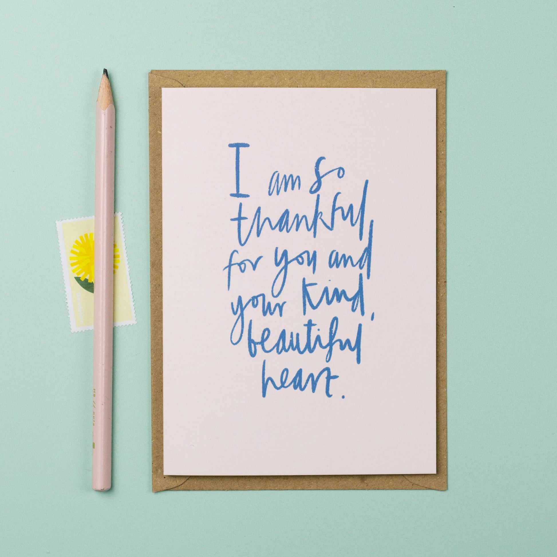 Greeting card with blue text on a light pink background, next to a pink pencil and small yellow card with a daisy on a light green background.