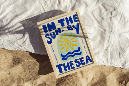 Poster with 'In the Sun, By the Sea' text and sun graphic on sand and fabric background