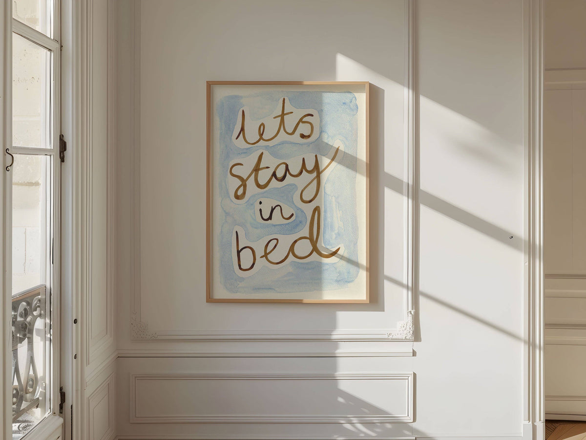 Framed artwork with 'let's stay in bed' text on a wall in a bright room.