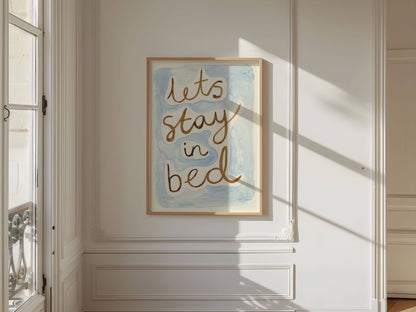 Framed artwork with 'let's stay in bed' text on a wall in a bright room.