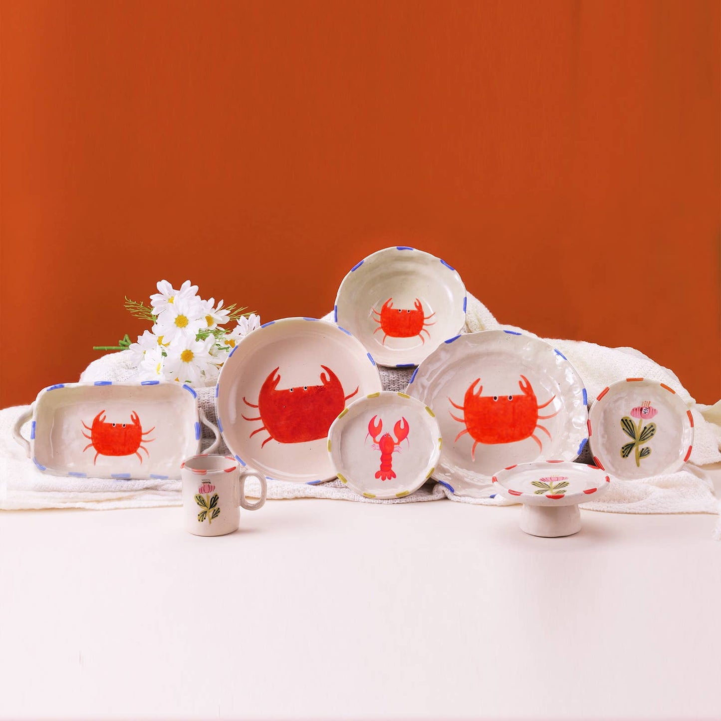 Set of ceramic plates and mugs with red crab and lobster designs on a white tablecloth against an orange wall.