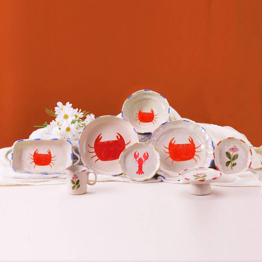 Set of ceramic plates and mugs with red crab and lobster designs on a white tablecloth against an orange wall.
