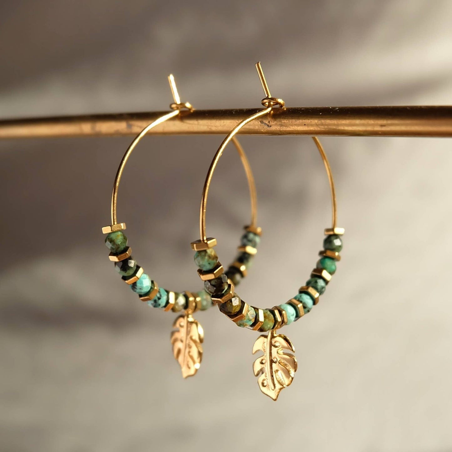 Gold hoop earrings with green beads and leaf charms on a neutral background