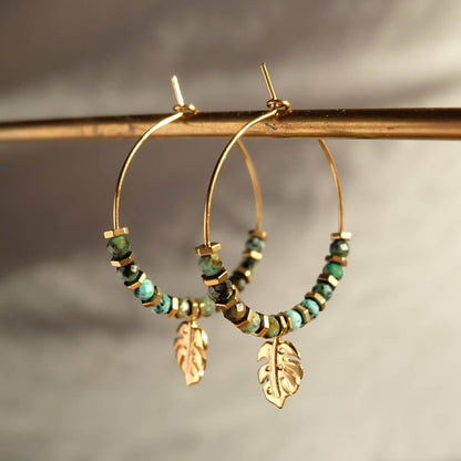Gold hoop earrings with green beads and leaf charms on a neutral background