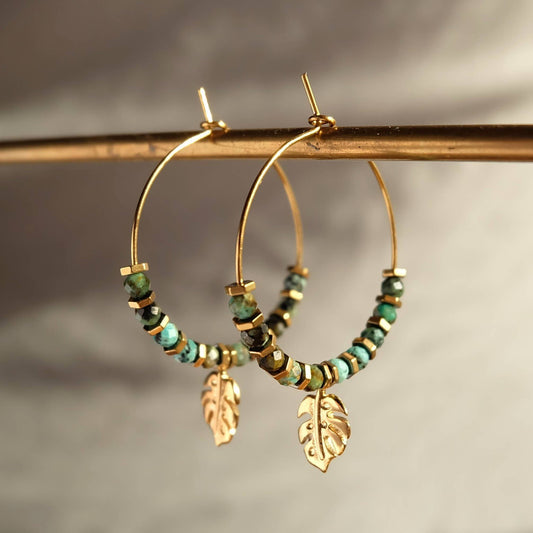 Gold hoop earrings with green beads and leaf charms on a neutral background