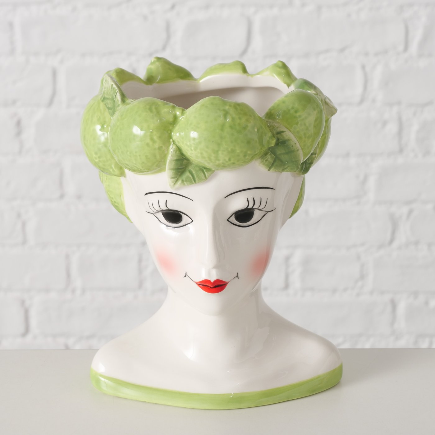 Ceramic vase shaped like a woman's head with green limes on her head against a white textured wall.
