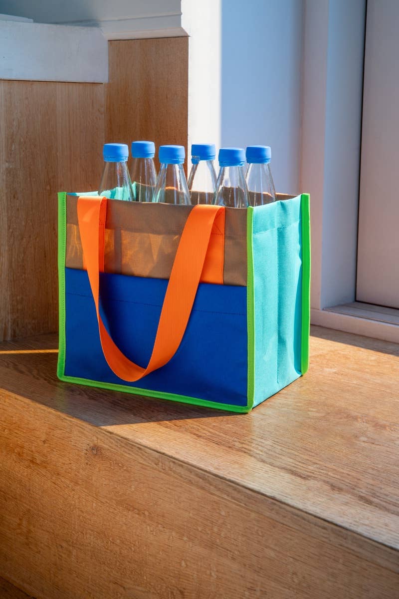Colorful tote bag with orange handles and blue base containing bottles on a wooden surface.