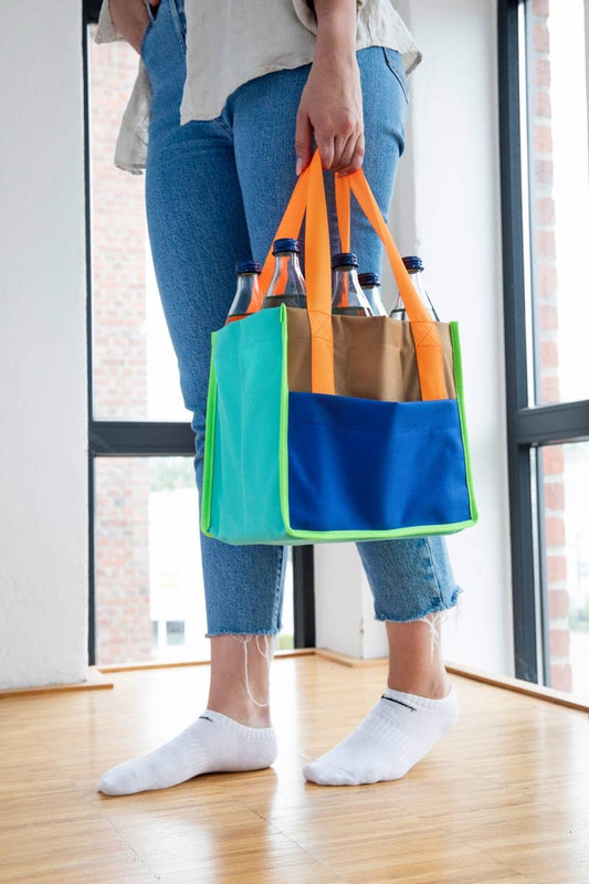 Person holding a colorful tote bag with bottles inside, standing in a room with large windows.