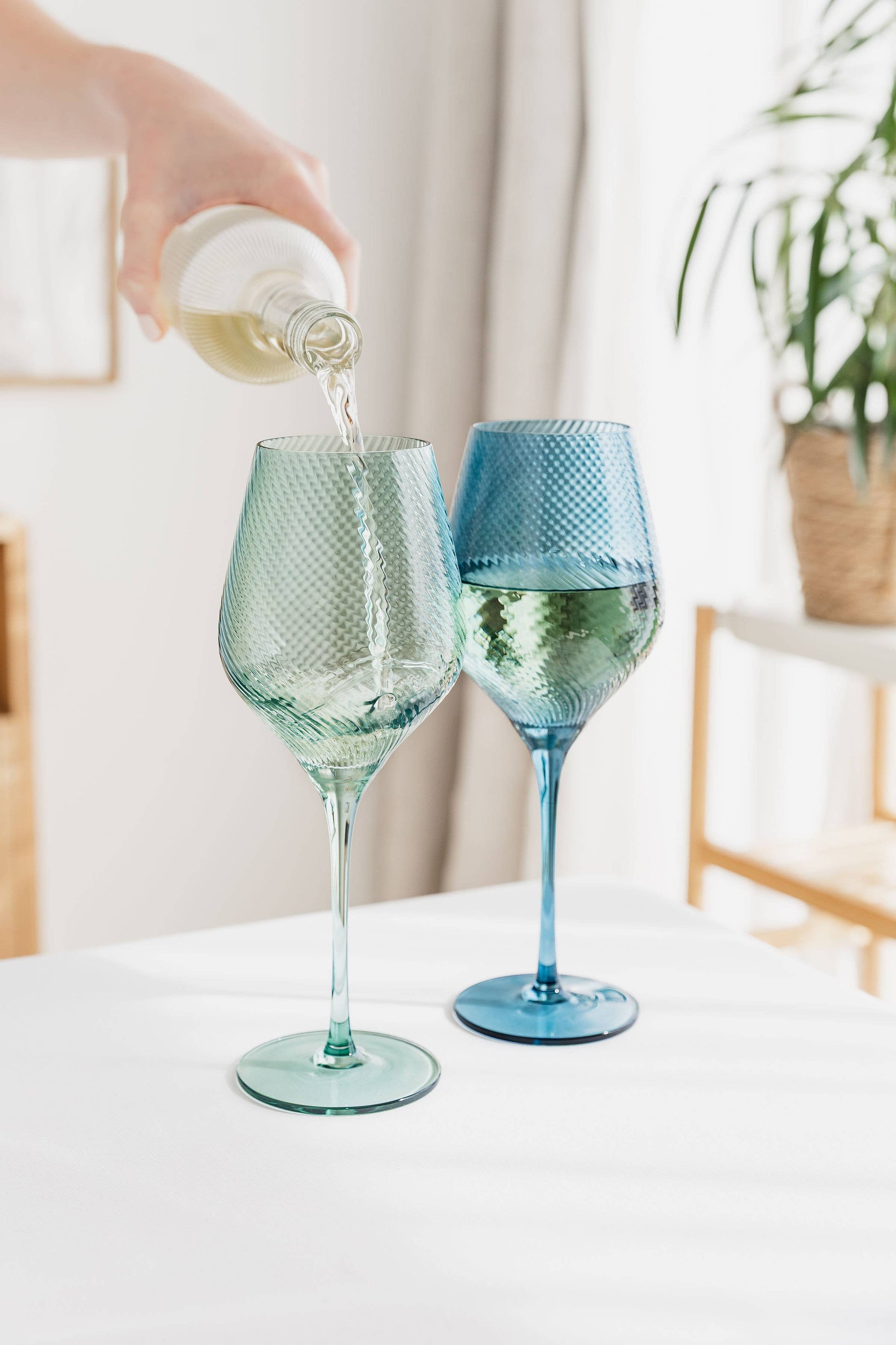 One blue, one green wine glasses being filled with white wine on a light surface.