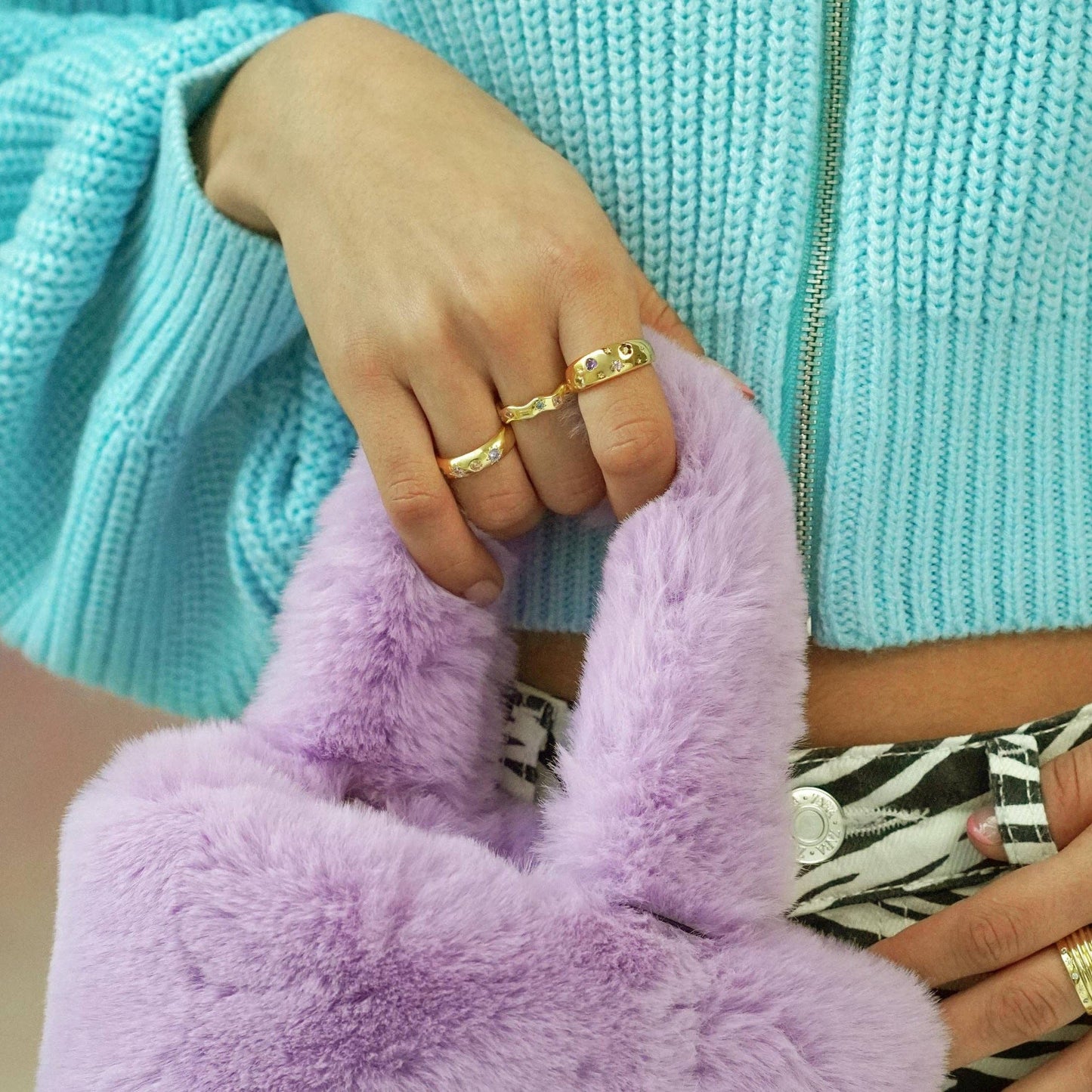 Person wearing gold rings holding a purple fluffy bag with a turquoise sweater in the background