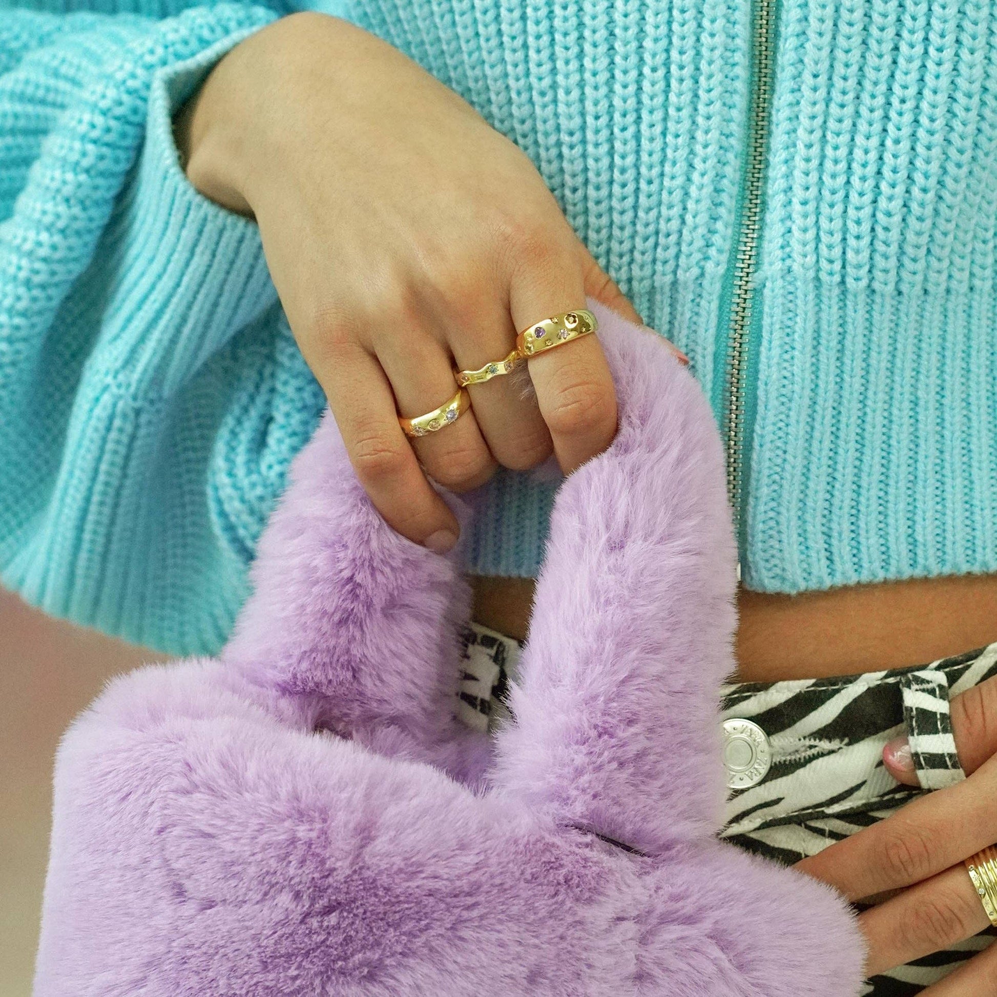 Person wearing gold rings holding a purple fluffy bag with a turquoise sweater in the background