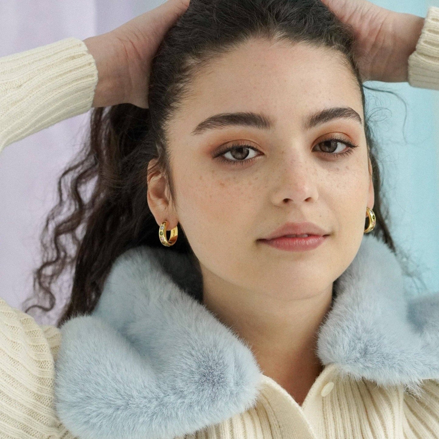 Woman wearing gold earrings with a light blue fur collar and beige sweater.