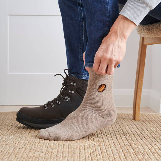 Person wearing brown hiking boots and beige socks with a brand logo on a carpeted floor.