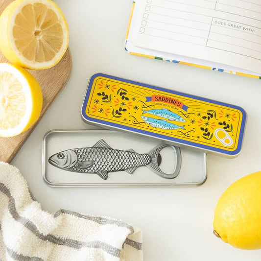Metallic fish-shaped bottle opener with decorative lid on a white surface with lemons and a towel.